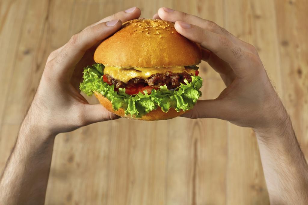 Eating Fast Food. Close Up Of Man's Hands Holding Delicious Classic American Hamburger Stacked With Beef Patty, Cheese, Lettuce, Onion And Tomato On Fresh Bun With Sesame. Point Of View. Nutrition